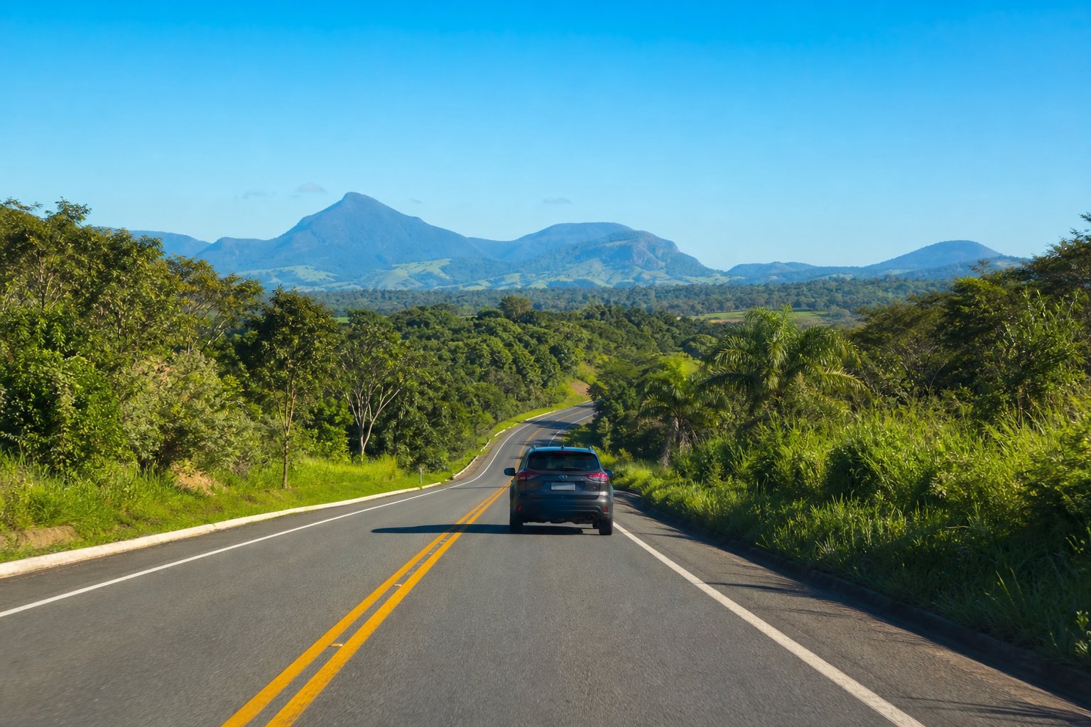 Carro moderno dirigindo por uma estrada sinuosa cercada por vegetação tropical verde e colinas ao fundo sob céu azul claro.